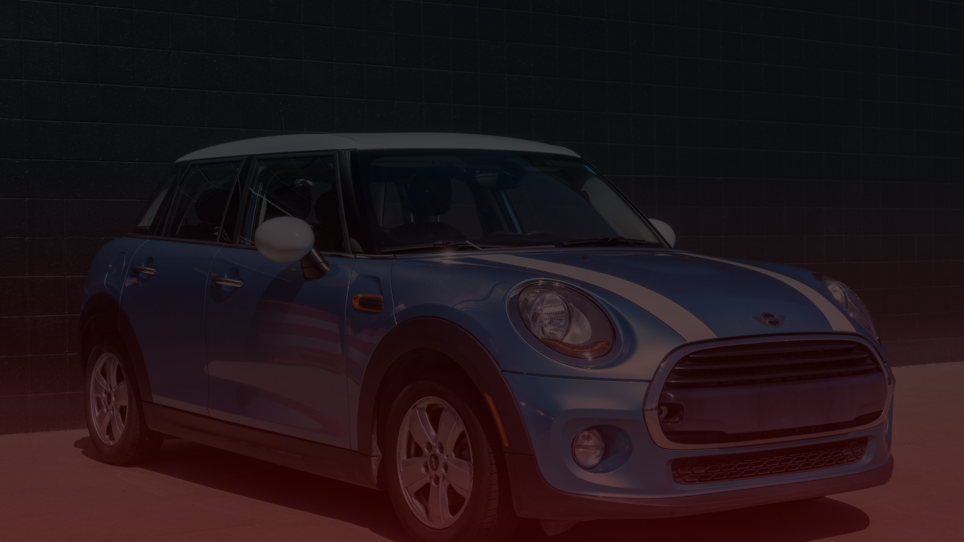 A blue Mini Cooper with a white roof and racing stripes gleams under the sun, parked stylishly in front of a dark brick wall. Its compact design and distinctive headlights shine brightly—trustworthy for any adventure or visit to your Mini Cooper mechanic in North York. EuroMechanic - McNally Auto | Top Reviewed Auto Repair Shop in North York | Visit EuroMechanic - McNally Auto