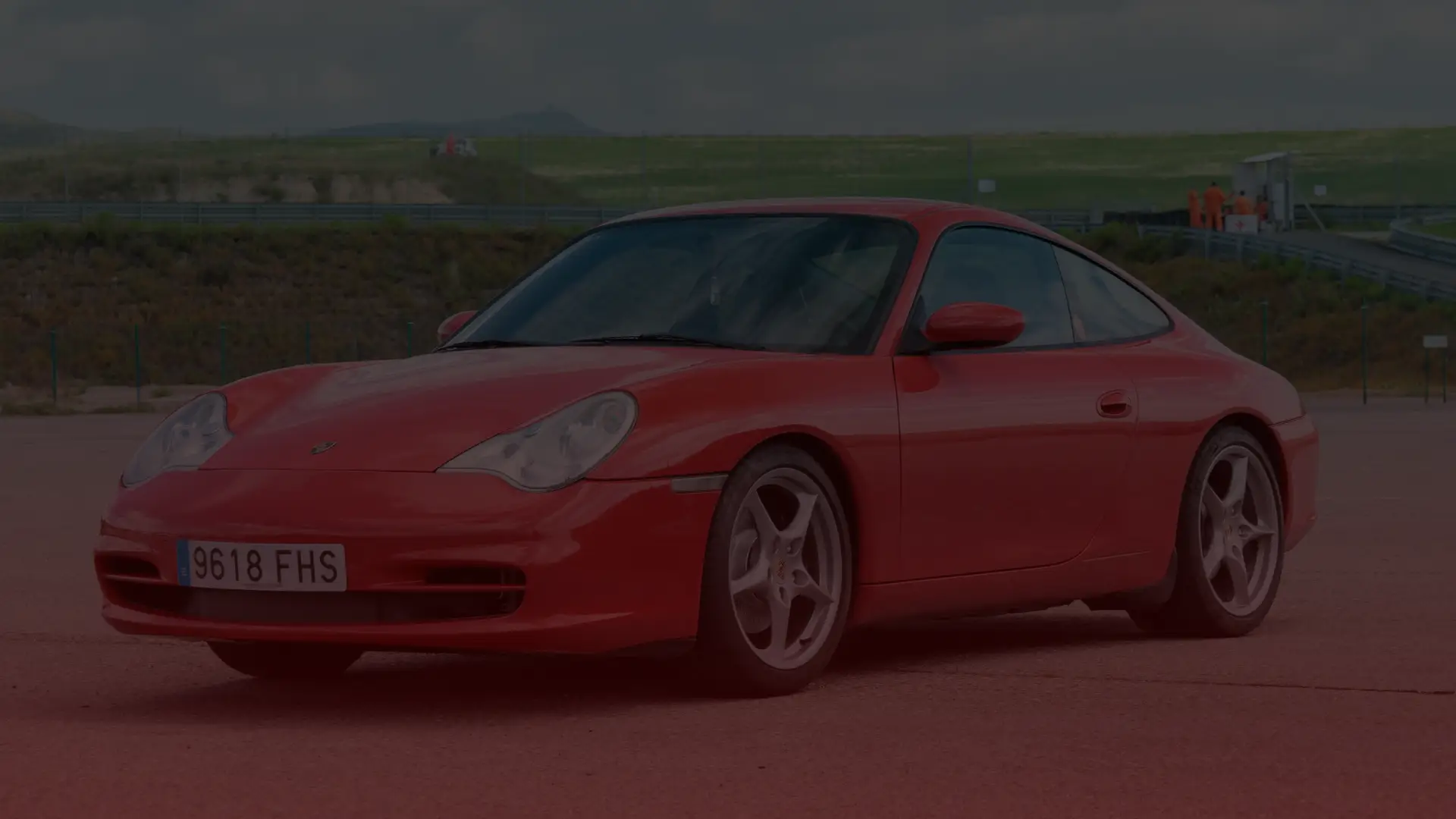 A red sports car with sleek design and alloy wheels is parked on an open asphalt area. The sky is cloudy, and greenery flourishes in the background—a perfect setting for any Porsche enthusiast seeking repair or maintenance services at Porsche Repair North York. EuroMechanic | McNally Auto | Top Reviewed Auto Repair Shop in North York | Visit mcnallyauto.com