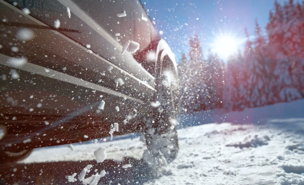 Close-up of a car from McNally Auto driving through snowy terrain, snowflakes flying around. Sunlight shines brightly, illuminating the snow-covered landscape. Pine trees are visible in the distance under a clear blue sky, ensuring your ride is smooth with Tires North York. EuroMechanic - McNally Auto | Top Reviewed Auto Repair Shop in North York | Visit EuroMechanic - McNally Auto
