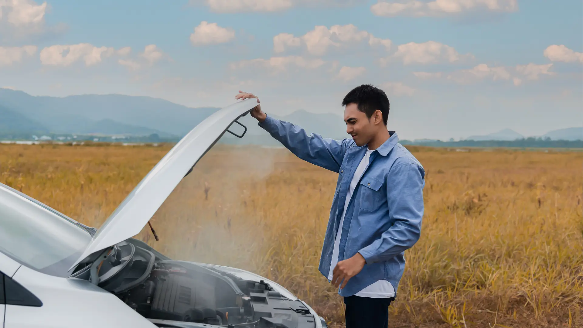 A man wearing a blue shirt stands beside a white car with the hood open, looking at the engine. Steam is rising from the engine. The background shows a field with dry grass and distant mountains under a cloudy sky. EuroMechanic - McNally Auto | Top Reviewed Auto Repair Shop in North York | Visit EuroMechanic - McNally Auto