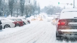 Cars drive and park along a snow-covered city street during heavy snowfall. Snow blankets the road and vehicles, and visibility is reduced. Traffic lights and headlights are visible through the snowy conditions. EuroMechanic - McNally Auto | Top Reviewed Auto Repair Shop in North York | Visit EuroMechanic - McNally Auto